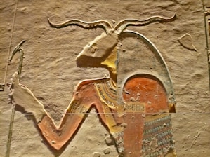 An ancient Egyptian carving featuring a prominent figure with an animal head resembling that of a goat or ram. The figure is adorned with traditional Egyptian attire, emphasizing colorful patterns and hieroglyphic designs. The carving is detailed, showcasing intricate designs on both the clothing and headgear against a textured stone background.