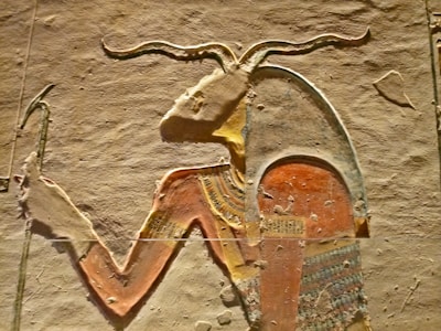 An ancient Egyptian carving featuring a prominent figure with an animal head resembling that of a goat or ram. The figure is adorned with traditional Egyptian attire, emphasizing colorful patterns and hieroglyphic designs. The carving is detailed, showcasing intricate designs on both the clothing and headgear against a textured stone background.