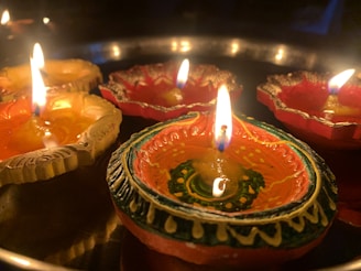 Decorative clay oil lamps, known as diyas, are arranged in a circular pattern with bright flames. The lamps are intricately painted with vibrant designs and colors, creating a warm and festive atmosphere. The glow of the flames casts a soft light over the scene.