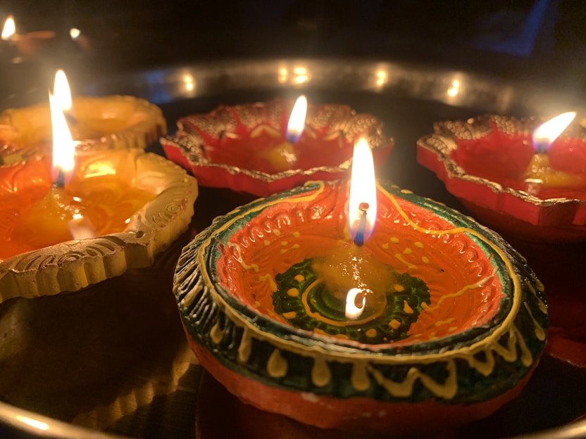 Decorative clay oil lamps, known as diyas, are arranged in a circular pattern with bright flames. The lamps are intricately painted with vibrant designs and colors, creating a warm and festive atmosphere. The glow of the flames casts a soft light over the scene.