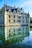A historic château with pointed turrets and elaborate stonework stands majestically by a body of water, creating a symmetrical reflection. The building features numerous windows and ornamented spires on its roof, and the calm water below mirrors its details. Bright sunlight illuminates the scene, casting light on the architectural features and creating a serene atmosphere.