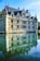 A historic château with pointed turrets and elaborate stonework stands majestically by a body of water, creating a symmetrical reflection. The building features numerous windows and ornamented spires on its roof, and the calm water below mirrors its details. Bright sunlight illuminates the scene, casting light on the architectural features and creating a serene atmosphere.