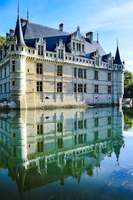 A historic ch&acirc;teau with pointed turrets and elaborate stonework stands majestically by a body of water, creating a symmetrical reflection. The building features numerous windows and ornamented spires on its roof, and the calm water below mirrors its details. Bright sunlight illuminates the scene, casting light on the architectural features and creating a serene atmosphere.