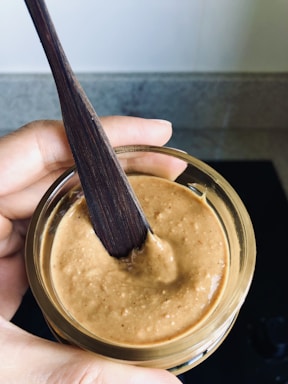 A hand holding a jar filled with a smooth, beige-brown creamy substance, likely peanut butter. A dark wooden spoon or spatula is dipped into the mixture. The background is a neutral-toned kitchen countertop.