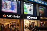 A series of shop signs and storefronts are illuminated at night. The stores include Monark and Equator, with mannequins and clothing visible inside. Above the stores, there are large advertisements featuring animated characters and children.