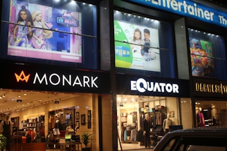 A series of shop signs and storefronts are illuminated at night. The stores include Monark and Equator, with mannequins and clothing visible inside. Above the stores, there are large advertisements featuring animated characters and children.