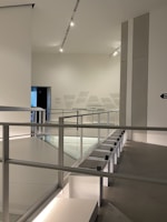 An interior space with modern architecture, featuring clean lines and minimalistic design. The area includes a railing alongside a path, illuminated by overhead lighting. The walls are adorned with subtle text or art, adding to the contemporary feel.