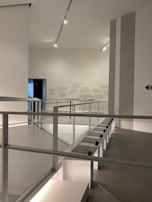 An interior space with modern architecture, featuring clean lines and minimalistic design. The area includes a railing alongside a path, illuminated by overhead lighting. The walls are adorned with subtle text or art, adding to the contemporary feel.