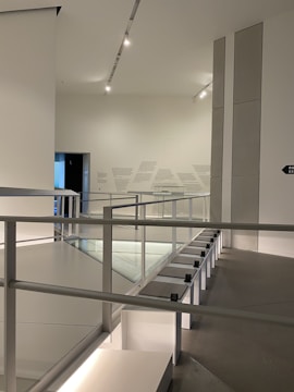 An interior space with modern architecture, featuring clean lines and minimalistic design. The area includes a railing alongside a path, illuminated by overhead lighting. The walls are adorned with subtle text or art, adding to the contemporary feel.