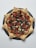 A star-shaped pizza with a unique crust design, topped with a variety of colorful vegetables including cherry tomatoes, black olives, and bell peppers. The pizza sits on a dark, round slate board, contrasting with the light background.