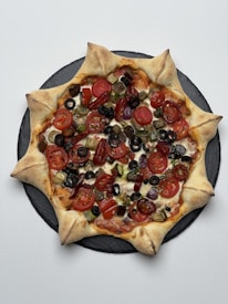 A star-shaped pizza with a unique crust design, topped with a variety of colorful vegetables including cherry tomatoes, black olives, and bell peppers. The pizza sits on a dark, round slate board, contrasting with the light background.
