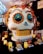 A large decorative skull with intricate designs stands on display, adorned with colorful flowers and feathers. Its eyes are detailed with bright blue and orange patterns, while the teeth are stylized in a golden hue. Surrounding the skull are decorative candles and smaller ornamental skulls, each housed in transparent boxes. The setting suggests a festive or celebratory theme.