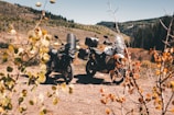 Two adventure motorcycles parked on a dirt path surrounded by autumn foliage and rolling hills under a clear blue sky. The motorcycles are equipped with gear, indicating a potential journey or trip. In the background, the landscape features a mix of greenery and patches of dried grass or bushes.