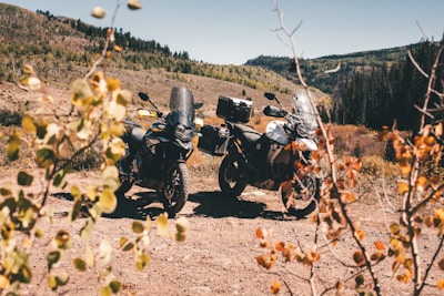 Two adventure motorcycles parked on a dirt path surrounded by autumn foliage and rolling hills under a clear blue sky. The motorcycles are equipped with gear, indicating a potential journey or trip. In the background, the landscape features a mix of greenery and patches of dried grass or bushes.