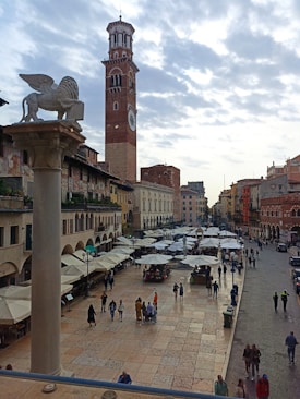 A bustling town square filled with people walking and socializing. Numerous market stalls with white canopies line the central area, while a tall brick clock tower stands prominently in the background. Ornate buildings with arched windows and columns surround the square, and a winged lion statue atop a column is visible in the foreground.