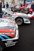 A collection of classic racing cars is displayed in an indoor exhibition space. Prominent sponsors' logos like Michelin and Martini are visible on the cars, which have a white body with red and blue stripes. There are a few people walking and admiring the vehicles. In the background, motorcycles and more cars can be seen.