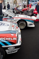 A collection of classic racing cars is displayed in an indoor exhibition space. Prominent sponsors' logos like Michelin and Martini are visible on the cars, which have a white body with red and blue stripes. There are a few people walking and admiring the vehicles. In the background, motorcycles and more cars can be seen.