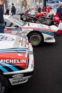 A collection of classic racing cars is displayed in an indoor exhibition space. Prominent sponsors' logos like Michelin and Martini are visible on the cars, which have a white body with red and blue stripes. There are a few people walking and admiring the vehicles. In the background, motorcycles and more cars can be seen.
