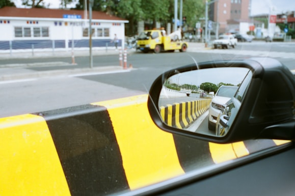 A side mirror of a car reflects another vehicle behind, set against a road with black and yellow striped barriers. A tow truck and other vehicles are seen on the street with a building featuring blue and white exterior walls in the background.