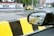 A side mirror of a car reflects another vehicle behind, set against a road with black and yellow striped barriers. A tow truck and other vehicles are seen on the street with a building featuring blue and white exterior walls in the background.