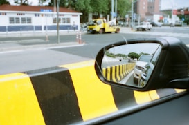 A side mirror of a car reflects another vehicle behind, set against a road with black and yellow striped barriers. A tow truck and other vehicles are seen on the street with a building featuring blue and white exterior walls in the background.