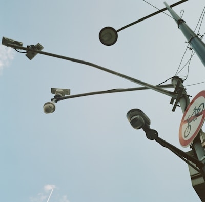 Several surveillance cameras are mounted on a tall metal pole, with long arms extending outward. Next to the cameras is a road sign indicating no bicycles. The sky is mostly clear with a few wispy clouds.