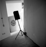 A black speaker on a tripod stand is positioned in the corner of a modern, minimalist space. The floor is tiled, and there are visible electrical outlets and cords running along the wall. In the background, an abstract circular artwork is mounted on a wall, with another circular window or patterned element nearby. The setting has a slightly tilted perspective, giving a dynamic feel.