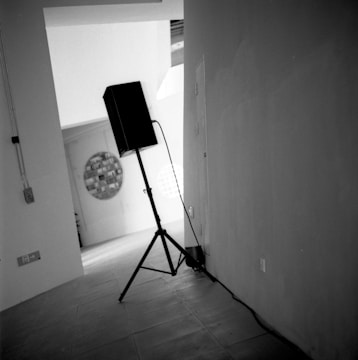 A black speaker on a tripod stand is positioned in the corner of a modern, minimalist space. The floor is tiled, and there are visible electrical outlets and cords running along the wall. In the background, an abstract circular artwork is mounted on a wall, with another circular window or patterned element nearby. The setting has a slightly tilted perspective, giving a dynamic feel.