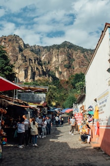 A lively market street is bustling with people walking along a cobblestone path. Shops and stalls line the sides, displaying various goods and colorful textiles. The background features majestic rocky mountains under a partly cloudy sky.