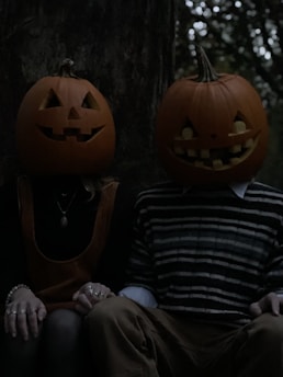 Two individuals sit side by side, each wearing a carved pumpkin with playful expressions as masks on their heads. Their hands are clasped together, and they are dressed in dark attire, creating a mysterious or eerie atmosphere. The background is dimly lit, resembling a forest setting.