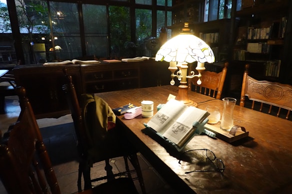 A cozy reading nook with a wooden table and chairs. A stained glass lamp illuminates an open book, glasses, a cup, and other small items on the table. Books are visible on shelves in the background, and there is a large window offering a view of greenery outside.