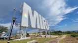 A large sign with the letters 'MIRI' is prominently displayed, set against a picturesque backdrop of a partly cloudy sky and lush greenery. The sign is supported by metal scaffolding, and there's a rainbow reflection visible on it. The area includes a path, a street lamp, and a parked vehicle.