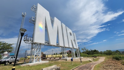 A large sign with the letters 'MIRI' is prominently displayed, set against a picturesque backdrop of a partly cloudy sky and lush greenery. The sign is supported by metal scaffolding, and there's a rainbow reflection visible on it. The area includes a path, a street lamp, and a parked vehicle.