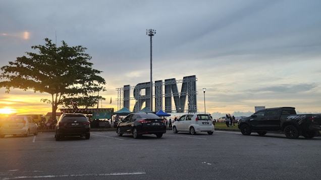 Cars parked in an outdoor area with a large metallic structure displaying the letters 'IRM' in the background. The setting sun and a tree add to the scenic view. A group of people are gathered near some tents.