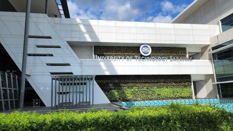 A modern building facade featuring a vertical garden with lush greenery, glass panels, and clean architectural lines. The sign reads 'University of Technology Sarawak,' situated above the greenery. The sky is clear with a few clouds visible.