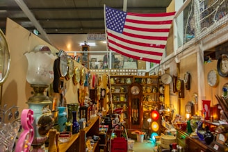 An indoor scene filled with an eclectic mix of vintage items, including clocks, lamps, and decorative objects. A large American flag hangs prominently on the wall. The shelves are cluttered with various collectibles such as ceramics and old records. The lighting is warm, creating a cozy atmosphere.
