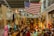 An indoor scene filled with an eclectic mix of vintage items, including clocks, lamps, and decorative objects. A large American flag hangs prominently on the wall. The shelves are cluttered with various collectibles such as ceramics and old records. The lighting is warm, creating a cozy atmosphere.