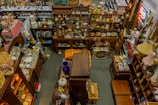 An overhead view of a cluttered antique store filled with various items. Shelves are stocked with vintage pottery, teapots, and glassware, while tables display decorative plates, vases, and lamps. There are wooden furniture pieces, small baskets, and books scattered throughout the space. A variety of eclectic and nostalgic objects create a sense of organized chaos.