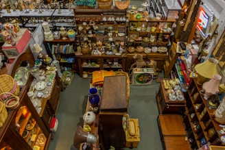 An overhead view of a cluttered antique store filled with various items. Shelves are stocked with vintage pottery, teapots, and glassware, while tables display decorative plates, vases, and lamps. There are wooden furniture pieces, small baskets, and books scattered throughout the space. A variety of eclectic and nostalgic objects create a sense of organized chaos.