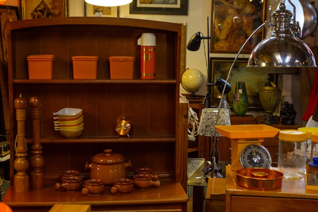 A cozy vintage kitchen setting with a wooden shelf that holds orange storage containers, a series of stacked bowls, a copper clock, a brown teapot, and smaller matching cups. Next to it, there are wooden pepper mills and a metal dome pendant light. An orange kitchen scale and assorted jars sit on the counter. In the background, there are various decorative items such as a globe, artwork, and vases.