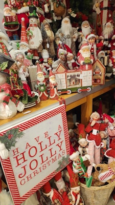 A festive display of Christmas-themed decorations, featuring an array of Santa Claus figures, snowmen, elves, and other holiday figurines. A bright sign with 'Have a Holly Jolly Christmas' is prominently placed in the foreground. The decorations are colorful and intricately detailed, with elements of greenery and rustic textures.