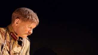 A person with short brown hair and a mustache, wearing a vintage military uniform with brass buttons and a red collar, gazes downward with a serious expression. The background is dark, highlighting the person's profile.