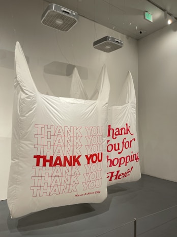 Large inflatable white bags are suspended from the ceiling in a room. The bags have printed messages in bold red letters, such as 'THANK YOU' and 'Thank You for Shopping Here!' The room's walls and ceiling are plain, and there are some ceiling fixtures visible.