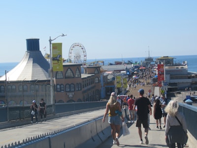 A bustling boardwalk filled with people walking towards a vibrant pier. The scene includes a large Ferris wheel in the background and various colorful buildings lining the path. The ocean is visible to the right, providing a scenic backdrop. The atmosphere is lively and vibrant, indicative of a popular tourist destination.