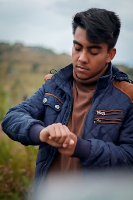 Man wearing a dark olive green tactical jacket checking his watch against a mountain backdrop.