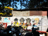 A vibrant piece of street art on a wall featuring cartoon-style characters, including monkeys wearing headphones, VR goggles, and a person holding an ice cream with playful expressions. The backdrop includes corrugated metal sheets and several parked motorcycles, with overhead trees casting shadows.