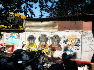 A vibrant piece of street art on a wall featuring cartoon-style characters, including monkeys wearing headphones, VR goggles, and a person holding an ice cream with playful expressions. The backdrop includes corrugated metal sheets and several parked motorcycles, with overhead trees casting shadows.