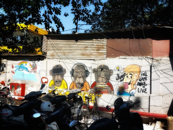 A vibrant piece of street art on a wall featuring cartoon-style characters, including monkeys wearing headphones, VR goggles, and a person holding an ice cream with playful expressions. The backdrop includes corrugated metal sheets and several parked motorcycles, with overhead trees casting shadows.
