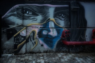 A detailed mural painted on a wall features a stylized human face with intricate line work and bold colors. The artwork incorporates elements of surrealism, combining abstract shapes and realistic eyes. The face appears to be divided with various colors, including pink, blue, and yellow. Some mechanical elements like red pipes and electrical boxes are integrated into the composition, adding an industrial touch.