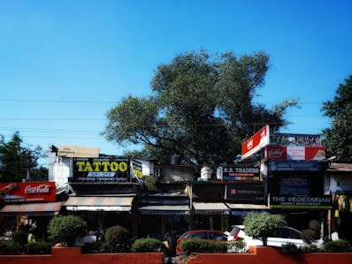 A street scene with multiple shops and signage, including a tattoo parlor offering discounts, a Coca-Cola advertisement, and various other small businesses. The area is lined with trees and shrubs, and there are parked vehicles in front.